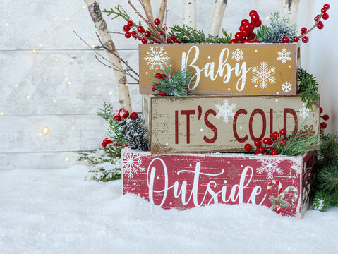 Background for personalized Christmas card. Country stacked crates on snow with text Baby It’s Cold Outside, birch branches and fairy lights, greenery and red berries on rustic white wood plank