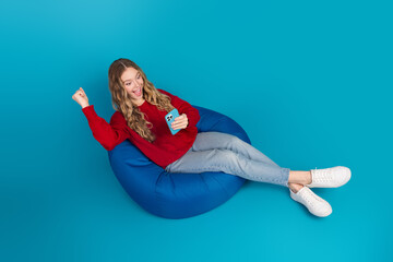 Cheerful young woman in red sweater relaxing on a comfortable beanbag while using her smartphone...