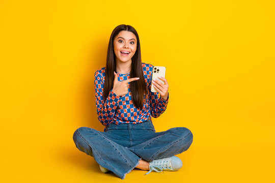 Young woman sits cross legged on bright yellow background smiling while showing smartphone and pointing at screen to invite engagement