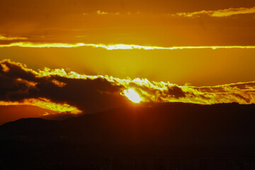 A powerful sunset paints the sky in glowing gold and deep orange hues over the dark silhouettes of hills.