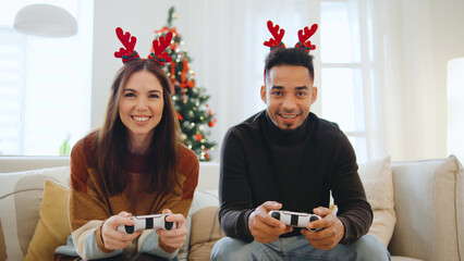 A cheerful couple wears reindeer antlers while playing video games together in their living room,...