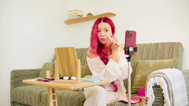 A woman with vibrant pink hair is applying makeup in her living room. She is filming a tutorial using a smartphone set on a tripod. The space is bright and inviting.