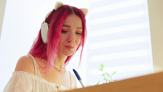 Young woman with pink hair wearing cat headphones, focused on writing at a stylish desk in a well-lit modern room during the day