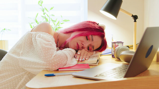 Student with pink hair rests while studying at home during the afternoon, with a laptop and notes scattered around her clean workspace