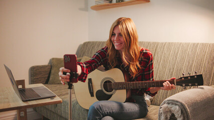 Red-haired young woman in red checkered shirt broadcasts online for blog and plays guitar