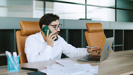 Business professional engaged in a phone call while working on a laptop in a modern office setting during daylight hours