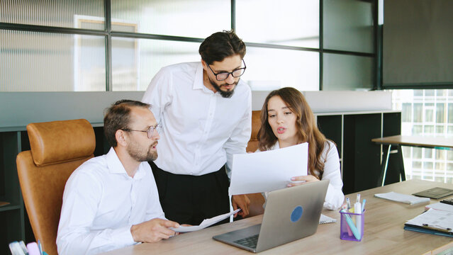 Three colleagues collaborate on a project, reviewing documents together at a well-organized desk in a contemporary office environment.
