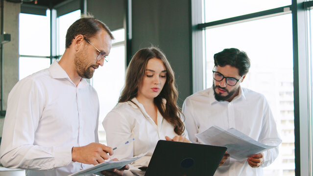 Three professionals review project documents and data on a laptop while collaborating in a bright office setting, sharing ideas and insights.