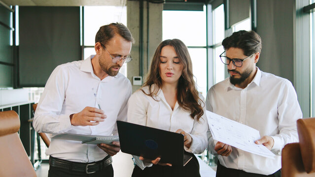 Three professionals review project documents and data on a laptop while collaborating in a bright office setting, sharing ideas and insights.