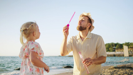 On a sunny beach in the late afternoon, a father playfully blows bubbles for his young daughter, who watches in delight by the shore.