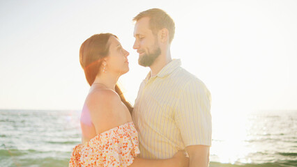 Fototapeta premium Two people stand close together at the beach, gazing into each other's eyes as the sun sets over the water, creating a warm and romantic atmosphere.