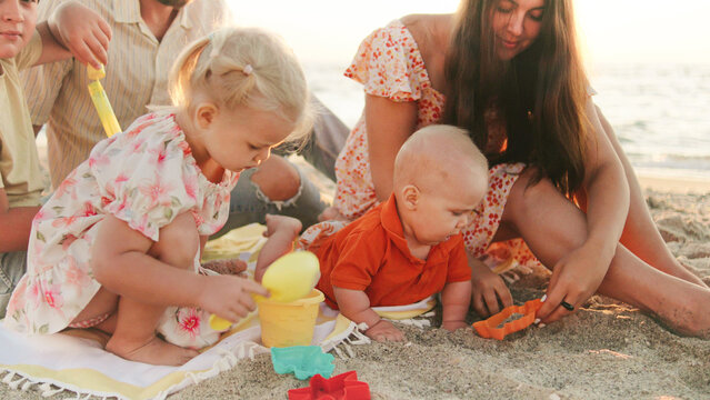 A family spends a joyful time on the beach as three young children play with colorful toys in the sand while the sun sets over the ocean.