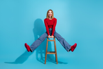 Lovely young woman in red shirt sitting on stool with cheerful pose against blue background...