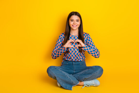 Bright young woman sits cross legged on a vibrant yellow background forming a heart with her hands for lifestyle fashion advertising and friendly branding