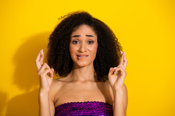 Young woman with curly hair crosses fingers in a bright yellow studio wearing a purple sequin top...