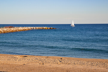 A tranquil seaside landscape with a sandy beach and a stone breakwater. A serene landscape with a yacht sailing on a calm sea