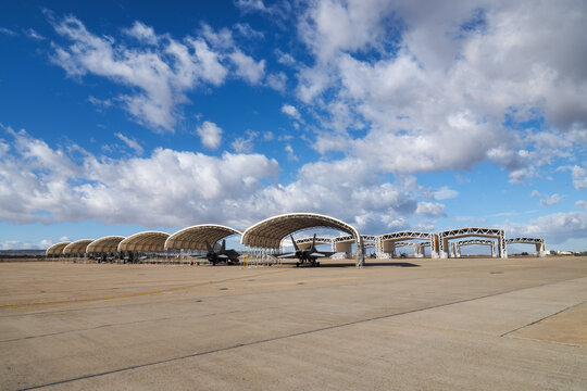 Row of twin engine multirole fighter jets parked under arched sun shelters on a concrete airbase apron under partly cloudy sky