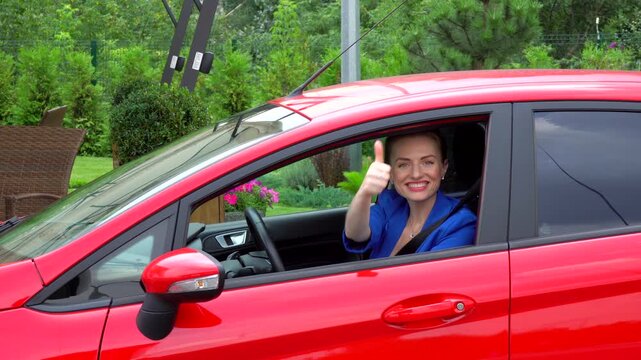 Young woman in blue suit drives red car. She stoppes and shows big thumb up. Young woman smiles and then drives away.