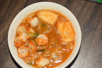 Spicy Indonesian seblak with chicken feet, tofu, sausage, crackers, lime, and vegetables in chili broth. Top view on white background.