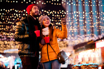 Joyful couple under festive lights enjoys warm drinks at night market celebrating christmas magic...