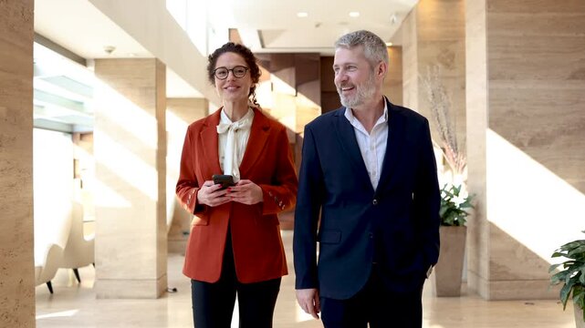 Two successful business partners, a mature man and a young woman, talking while walking through a bright and modern hotel or office lobby