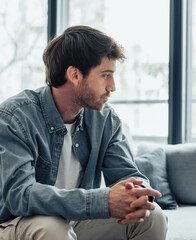 Thoughtful serious man sitting on sofa alone at home, lost in thoughts, thinking about problem...