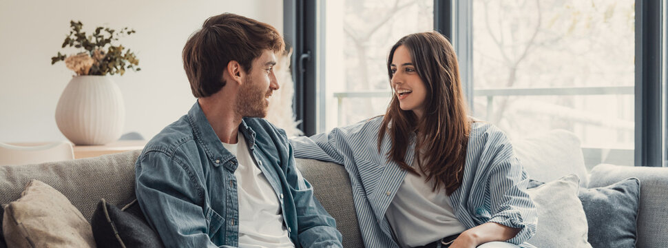 Young happy couple having fun talking laughing relaxing at home on couch, boyfriend embracing girlfriend telling funny joke sitting on sofa, humor in relationships, enjoying weekend together