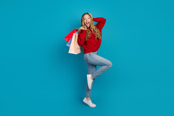 Pretty young woman in a red sweater holding shopping bags and posing happily against a vibrant blue...