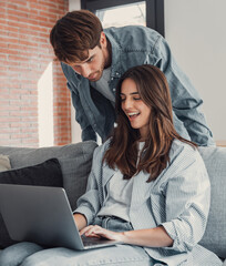 Happy millennial couple sit relax on couch in living room, smiling young husband standing from back...