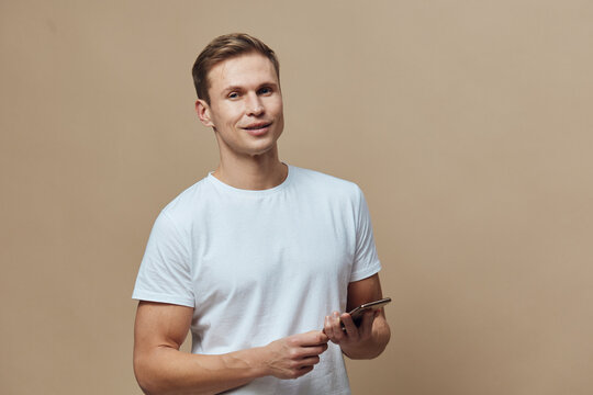 smiling young caucasian man hold smartphone use mobile phone casual white t-shirt beige background digital nomad concept - Powered by Adobe