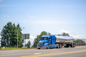 Bright blue big rig semi truck with low profile extended cab transporting cargo in cylinder bulk semi trailer running on the two lines city road