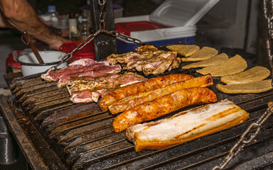 Grilled beef, pork belly and chorizos on barbecue with folded arepas and Colombian cook preparing food in outdoor cooking scene
