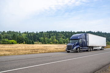 Dark blue big rig pro grade semi truck with dry van semi trailer running on the flat highway road with dry grass on the meadow on the side