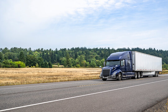 Dark blue big rig pro grade semi truck with dry van semi trailer running on the flat highway road with dry grass on the meadow on the side