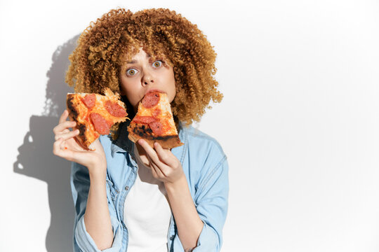 Surprised young woman with curly hair holding two slices of pizza, showcasing enjoyment and indulgence in a vibrant and playful manner