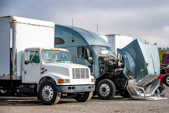 Gray broken big rig semi truck with open hood standing in row on the parking lot with another trucks waiting for repair and maintenance