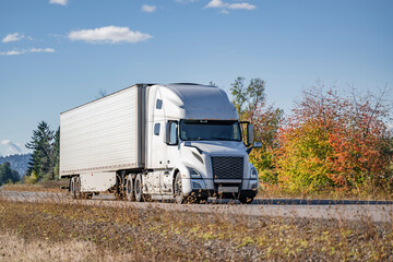 Industrial standard white big rig semi truck with extended cab transporting cargo in refrigerated semi trailer running on the autumn narrow road