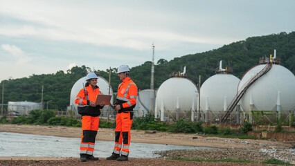Two engineers in orange safety gear and hard hats stand on a coastline. They are using a laptop and radio to inspect large industrial gas tanks in the background.