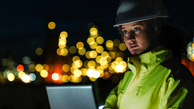 A close up of a female industrial worker in a hard hat and safety gear looking at a lit up factory or refinery at night. The background lights are blurred into bokeh.