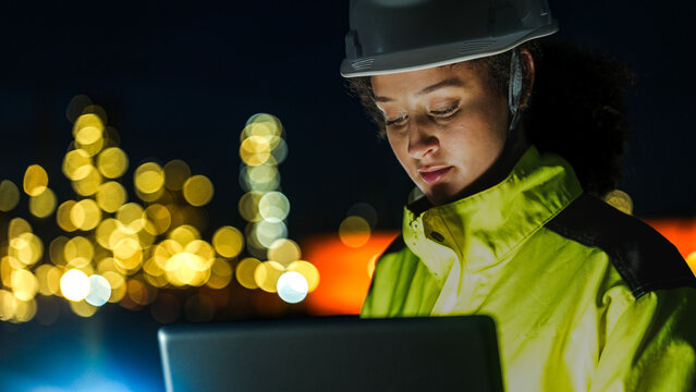 A petrochemical engineer in PPE monitors refinery operations during a night shift. She uses a laptop for real-time data analysis in front of lit-up processing infrastructure.