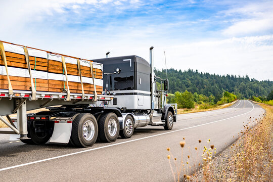 Powerful black and white classic American big rig semi truck tractor transporting fastened lumber wood on flat bed semi trailer driving on the summer winding road