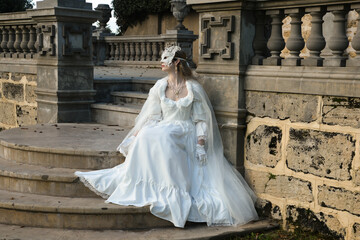 Obraz premium portrait of beautiful young blonde model wearing historical white wedding gown, fantasy cloak and butterfly masquerade mask. wandering ethereal figure in romantic fairytale castle garden background