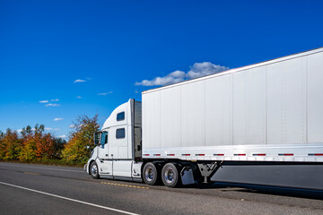 Pro white big rig semi truck tractor with high cab transporting cargo in dry van semi trailer running on the autumn highway road with yellow frees on the side