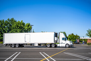Side view of the big rig white day cab semi truck with refrigerated semi trailer standing on the marked parking lot taking a break