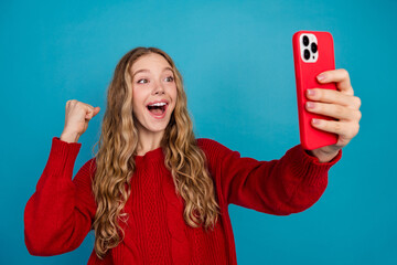 Adorable young woman taking cheerful selfie while celebrating with a smartphone in casual red...