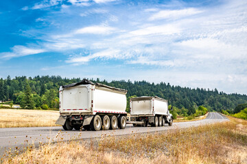 White day cab big rig tipper truck with two tip trailers running on the narrow road with meadows and forest on the hill
