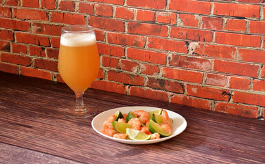 A plate of boiled shrimp with slices of ripe lime and a tall glass of wheat beer on a wooden table.