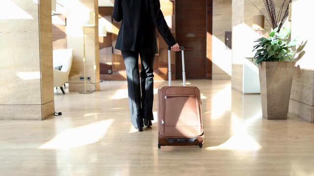 Unrecognizable businesswoman in a formal suit pulling a trolley bag. Rear view of a female traveler walking through a luxury hotel hall