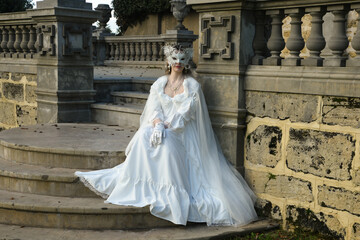 portrait of beautiful young blonde model wearing historical white wedding gown, fantasy cloak and butterfly masquerade mask. wandering ethereal figure in romantic fairytale castle garden background