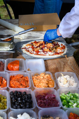 Chef placing fresh toppings on rolled out pizza dough on paddle to make individual wood fired pizza in food truck outdoors

